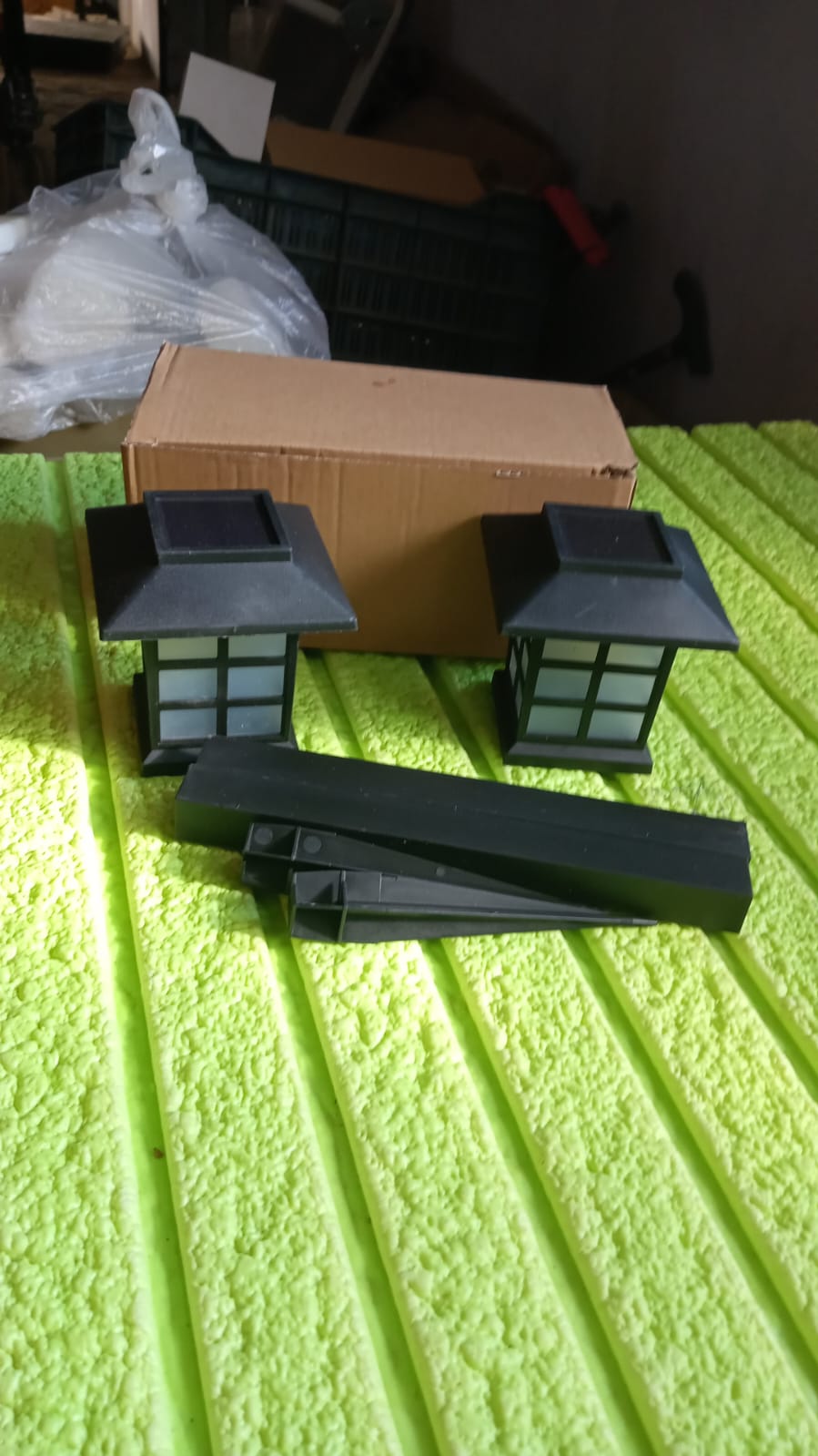 Two black solar light tops, several black pole pieces, and a brown cardboard box rest on a textured green surface.